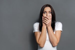 Keep silence. Scared woman covering mouth with hands while posing to camera on gray studio background. Shocked girl close lips with palms, speak no evil concept